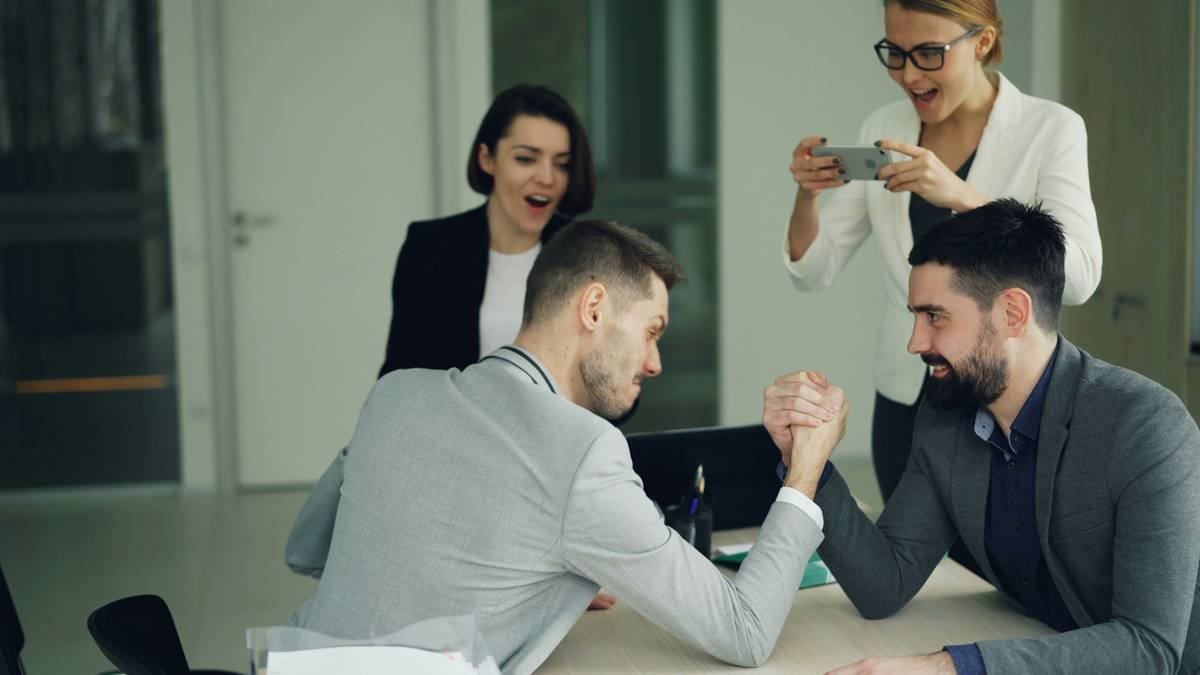 Male office workers are having fun doing arm-wrestling while cheerful girls are recording competition using smartphone and cheering guys. Work break and youth concept.