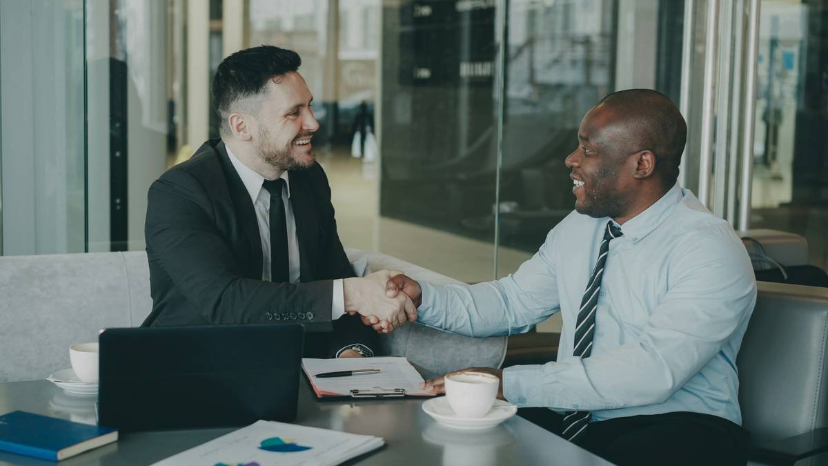 Two business colleagues discussing their business contract and looking at notepad in stylish cafe indoors. African American businessman puts his signature and shakes the hand of Caucasian man